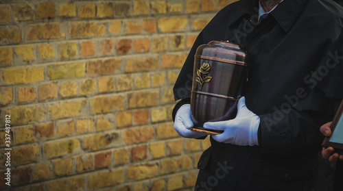 Funeral director or undertaker carrying a smooth metal urn in his hands, filled with ash towards the grave. Brick wall in the background. Funeral procession photo