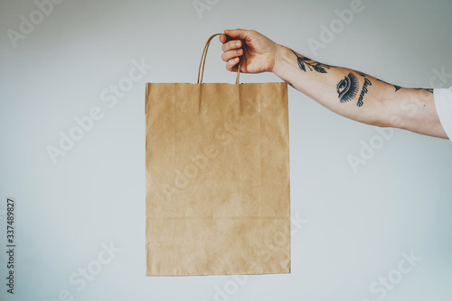 cropped shot on a hand with tattoos that holding craft paper package with empty space for your logo or design, mock-up of shopping bag with handles. White wall background