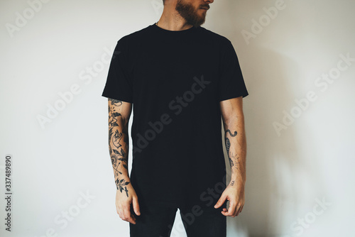 young attractive bearded man with tattoos, dressed in a black blank t-shirt, posing on a white wall background. Empty space for you logo or design.