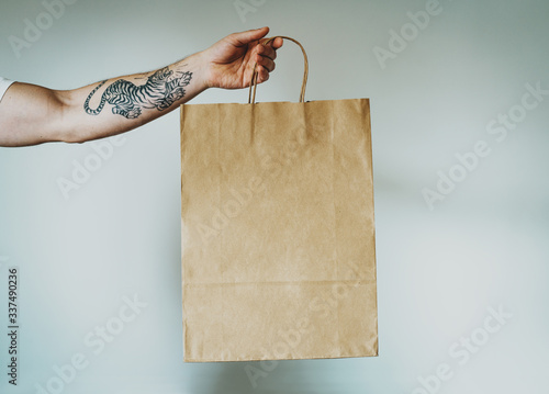 cropped photo on a hand with tattoos that holds craft paper package with empty space for your logo or design, mock-up of shopping bag with handles. White wall background photo