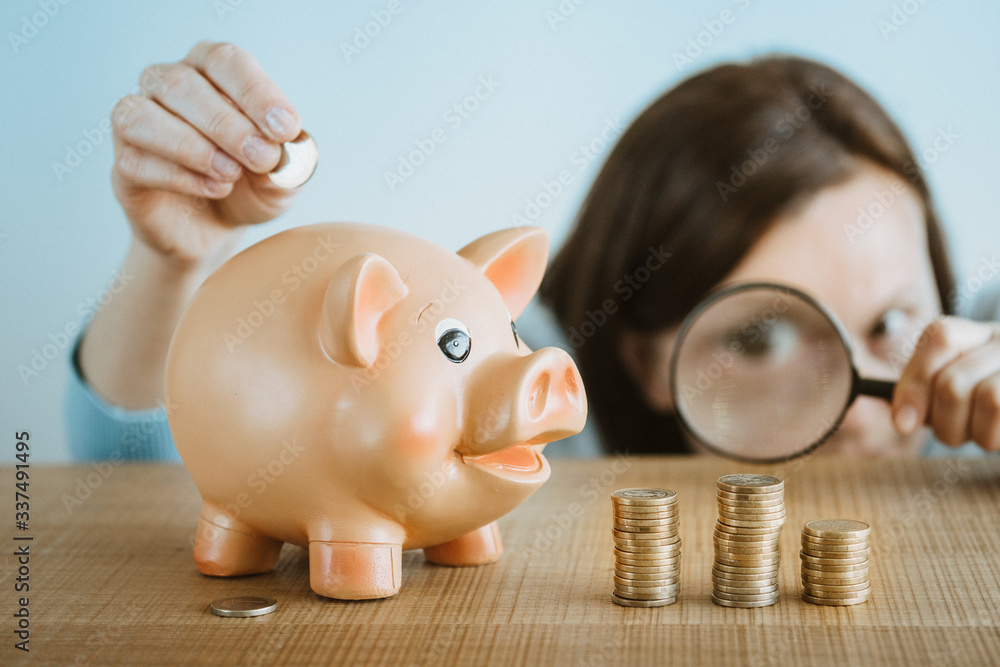 Young woman puts euro coin into a piggy bank and looks over the magnifying glass how coin tower grows. Investments and savings