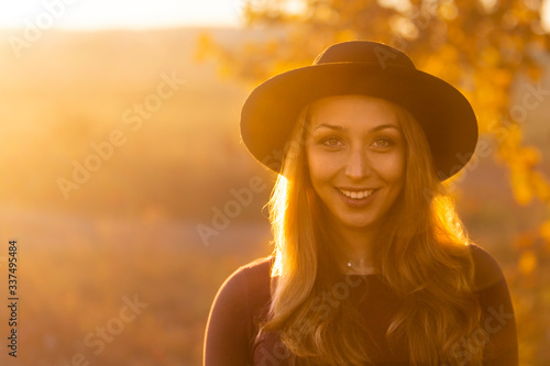 Happy beautiful caucasian girl with long hair in black hat smiles and looks at camera. Outdoors on warm sunny day. Nature yellow toned background. Enjoying life and nature. Fresh air, environment