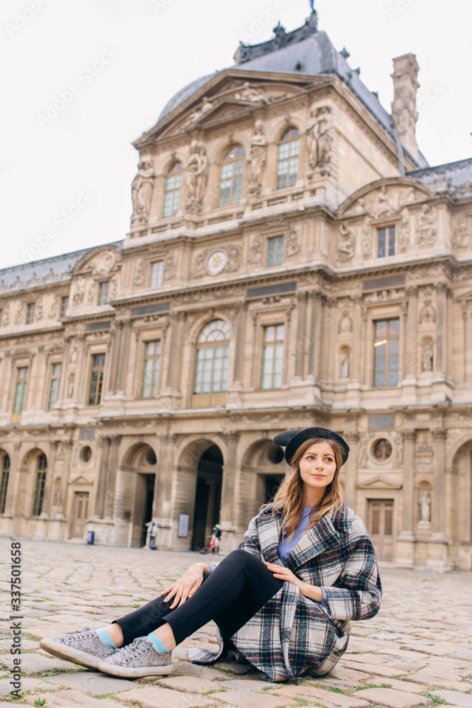 A girl in a beautiful coat stands near the Louvre.