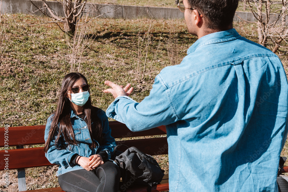 Man argues with his hands during a discussion with a woman sitting on a park bench wearing a face mask