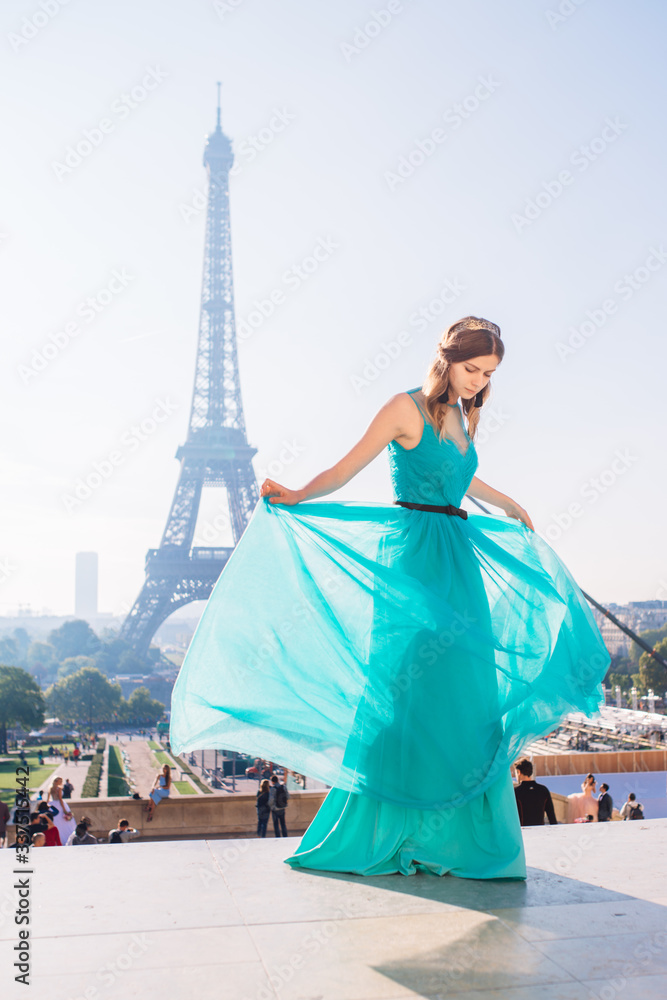 A young beautiful and elegant girl in a blue and green dress stands against the background of the Eiffel tower in Paris.