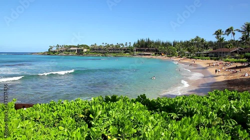 Napili Bay on Maui with aqua-blue water and gentle white waves rolling to the sandy shore with condominiums surrounding it on a bright and sunny Hawaii day.