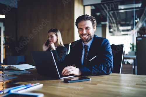 Portrait of cheerful male corporate director 30s sitting at office desk with laptop and smiling at camera, happy Caucasian businessman in elegant clothing enjoying work day in company firm