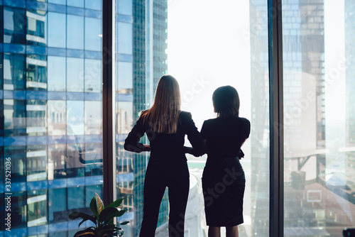 Back view of female colleagues in formal wear standing near window looking at modern exterior of skyscrapers in business center, prosperous women standing together planning prosperous future success © BullRun