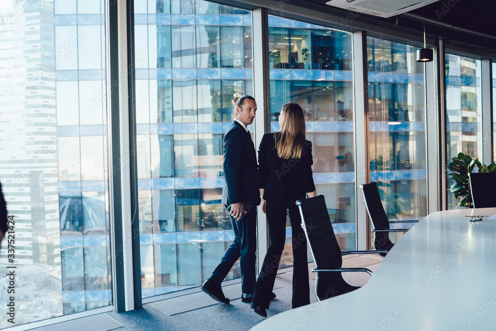 Fototapeta premium Male and female managers in formal outfit talking to each other strolling in office interior together, prosperous 30s woman and man business partners discussing planning and working process