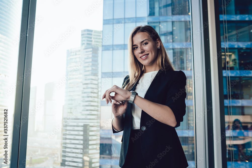 Cheerful prosperous blonde woman in elegant outfit looking at camera standing near window in office,caucasian female manager checking time on trendy wearable wrist watch satisfied with bracelet Cheerful prosperous blonde woman in elegant outfit looking at camera standing near window in office,caucasian female manager checking time on trendy wearable wrist watch satisfied with bracelet