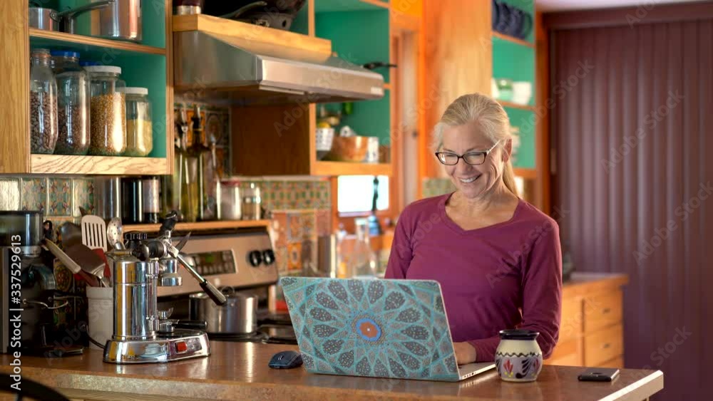 Talking to someone on her screen and smiling, a middle aged woman types on her keyboard taking notes while in the kitchen. Talking to someone on her screen and smiling, a middle aged woman types on her keyboard taking notes while in the kitchen.