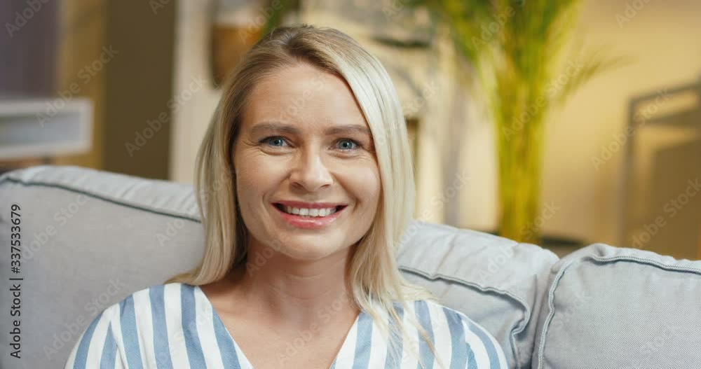 Close up portrait of Caucasian pretty smiling woman sitting on couch in room. Beautiful cheerful blonde female model with smile on face posing to camera at sofa in apartment inside. Indoor concept Close up portrait of Caucasian pretty smiling woman sitting on couch in room. Beautiful cheerful blonde female model with smile on face posing to camera at sofa in apartment inside. Indoor concept