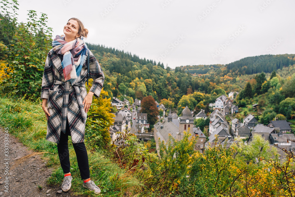 a girl in a coat stands against a panoramic view of Monschau, despite the wet weather.