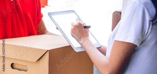 Young courier delivery man in red uniform holding a parcel cardboard box delivering package and woman putting signature in tablet to receiving package at home.online shopping and transport cargo 