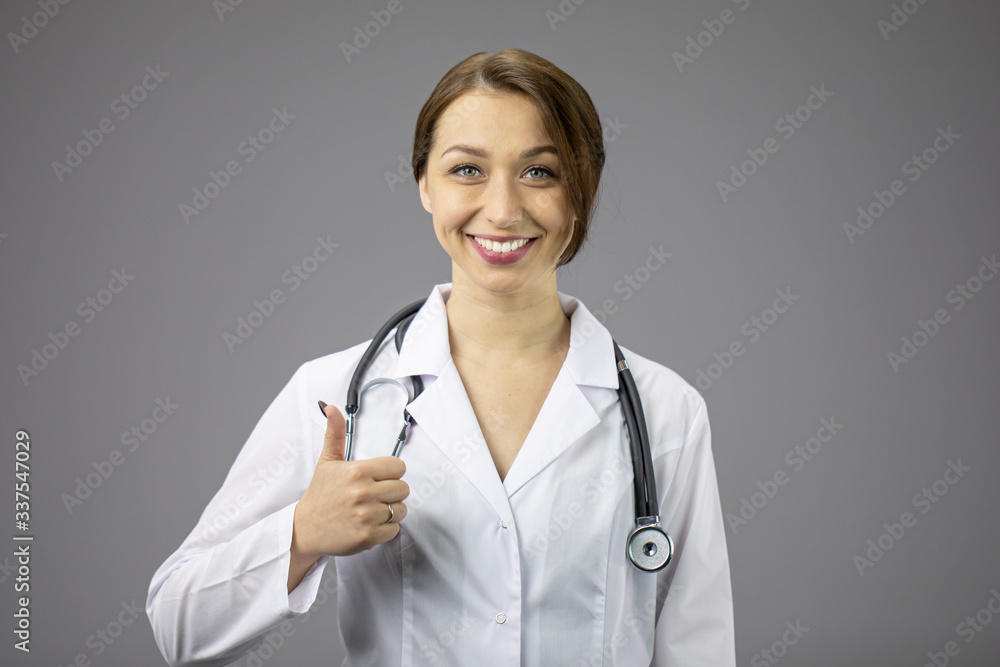 Portrait of an attractive female doctor in a white medical coat with a stethoscope on her neck. Thumb lifted upwards, clicks like. Snow-white smile. Red lips. Grey background. Studio shot.