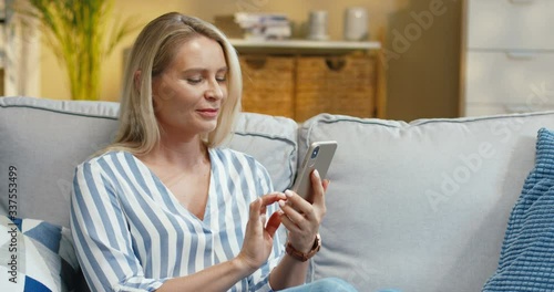 Portrait of charming dreamy blonde woman tapping on smartphone while resting on sofa in apartment in morning. Beautiful joyful Caucasian female writing message on cellphone indoor. Leisure concept photo