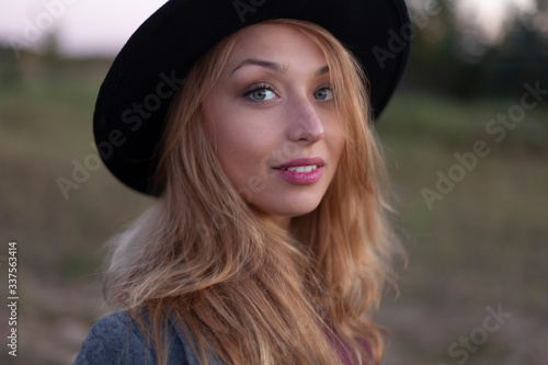 Close up portrait of beautiful caucasian woman with long fair hair in black hat looking at camera outside. Green nature backsides. Back to basics, relaxing concept © Model Republic