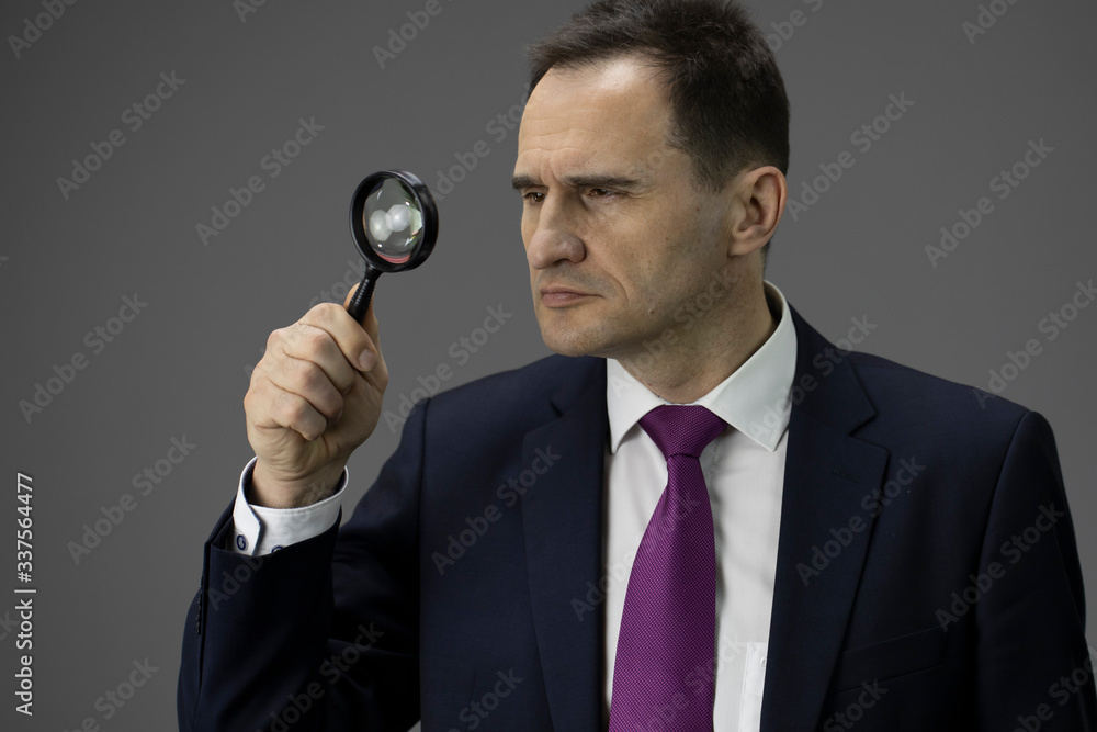 Concentrated male adult ceo in formal suit, tie looking through magnifier, isolated grey background. Copy space. Handsome man analysing business situation, reprofiling, search for new opportunities Concentrated male adult ceo in formal suit, tie looking through magnifier, isolated grey background. Copy space. Handsome man analysing business situation, reprofiling, search for new opportunities