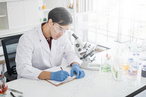 A male scientist with black hair wearing white coat and protective glassware writing on a clipboard next to a microscope in a laboratory setting with test tubes.