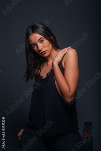 Sexy brunette wearing a black tank top and black pants posing while sitting on the top edge of a chair inside a studio on a dark background