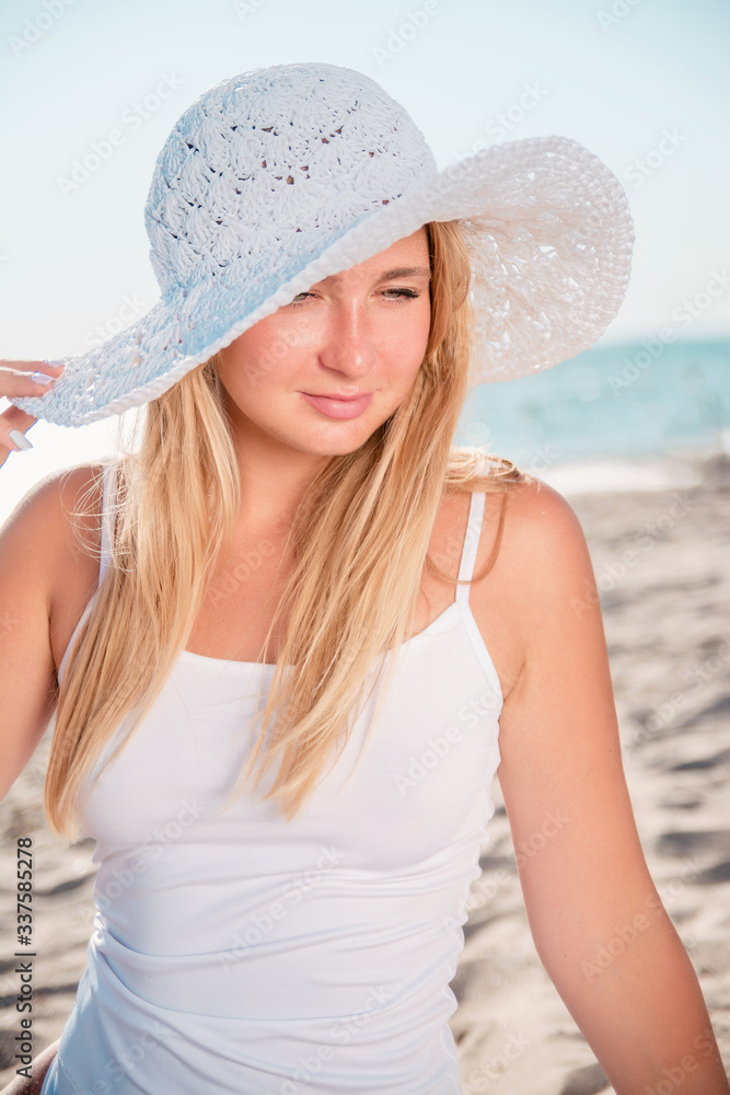 Woman in hat crouched on sand by sea. Portrait of a blonde girl in white sunbathing on a beach.
