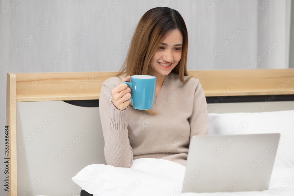 A beautiful Asian woman, Thai woman, is sitting and working with a notebook computer in the bed. The other hand is carrying coffee to drink to cure sleepiness in the bedroom in the apartment. A beautiful Asian woman, Thai woman, is sitting and working with a notebook computer in the bed. The other hand is carrying coffee to drink to cure sleepiness in the bedroom in the apartment.