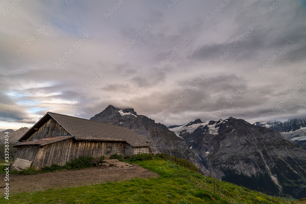 An alpine cabin hut on the edge of a grassy hill with the Wetterhorn and Schreckhorn mountain range in the background in Grindelwald Switzerland.