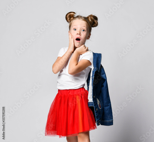 Shocked blonde kid girl with funny buns in red skirt and white t-shirt poses sideways to us holding hand at her cheek photo