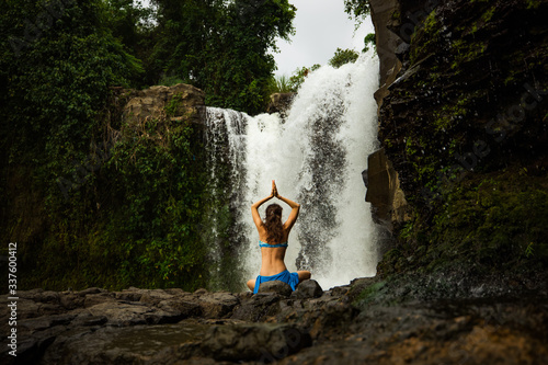 Young Caucasian woman sitting on the rock  meditating  practicing yoga at waterfall. Namaste mudra. View from back. Tegenungan waterfall in Ubud  Bali  Indonesia.