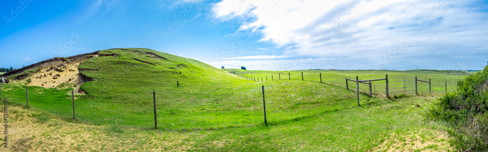 Wide panorama of green pastures with rolling hills and agricultural fence under beautiful blue sky