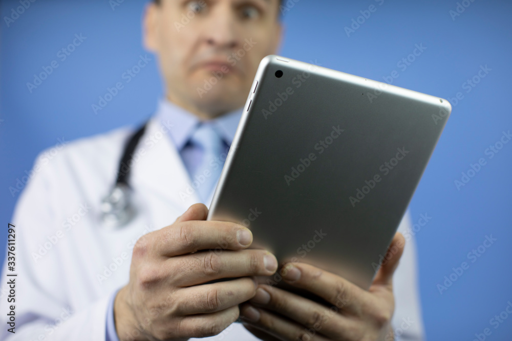 The doctor looks at the tablet with the results of the patient's tests. Adult male physician with stethoscope dressed in white medical coat holding a tablet. Blue background. The doctor looks at the tablet with the results of the patient's tests. Adult male physician with stethoscope dressed in white medical coat holding a tablet. Blue background.