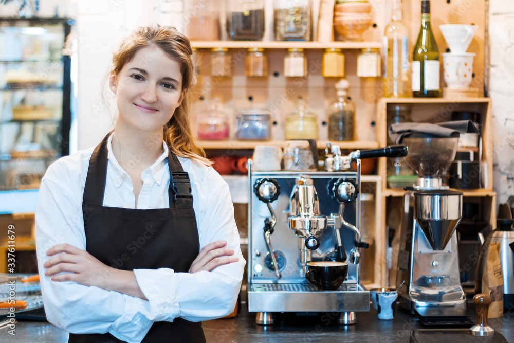 Young woman in brown apron is ready to take order from client customer in cafe, restaurant. Smiling barista on workplace. Professional coffee machine for preparing americano, espresso on background.