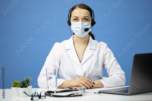 Remote medical consultations during quarantine and self-isolation. Online doctor consultation. Young female medical worker in white coat, mask and headset advises patients in clinic, copy space