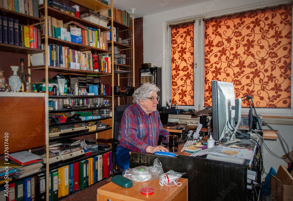 Elderly white haired man working at home on the desktop computer in a mess. Bookshelf with many books and antiviral masks ready Elderly white haired man working at home on the desktop computer in a mess. Bookshelf with many books and antiviral masks ready