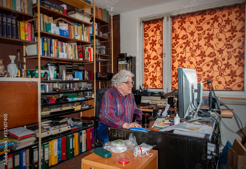 Elderly white haired man working at home on the desktop computer in a mess. Bookshelf with many books and antiviral masks ready