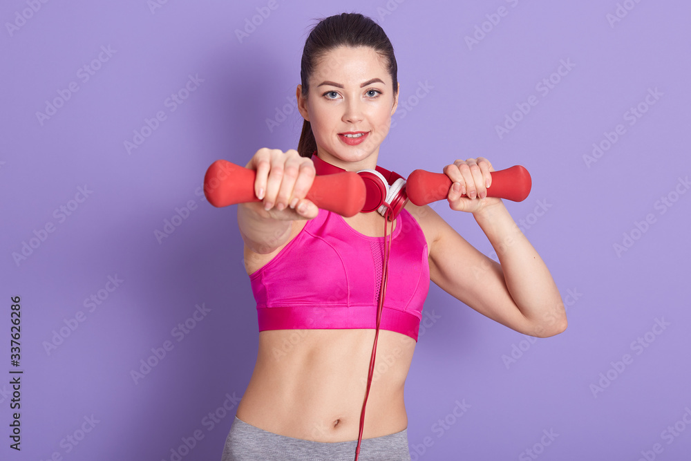 Indoor picture of sporty charismatic good looking female holding two red dumbbells, having headphones around neck, doing sports, having physical training, wearing sport clothes. Sport concept. Indoor picture of sporty charismatic good looking female holding two red dumbbells, having headphones around neck, doing sports, having physical training, wearing sport clothes. Sport concept.