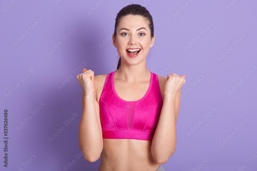 Horizontal shot of charming sportswoman posing with clenching fists isolated over lilac studio background and yelling something, being in good mood, happy with her result. sport and fitness concept.
