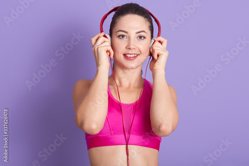 Closeup portrait of adorable woman training in gym with red headphones, sportswoman wearing pink sports bra, lady looking directly at camera and smiling, posing isolated over lilac studio background.