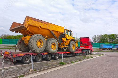 Big truck with a low platform trailer carrying a tipper truck on a public parking area of a truck stop.