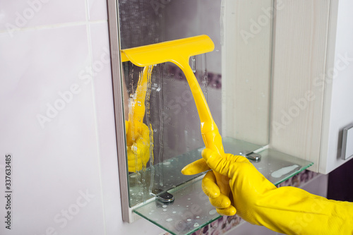  Cleaning the bathroom mirrors. A hand in a yellow glove washes a mirror with glass scraper.