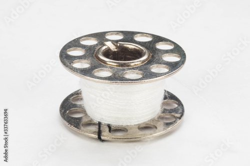 Close up of a metal sewing machine bobbin with white thread on a white background.