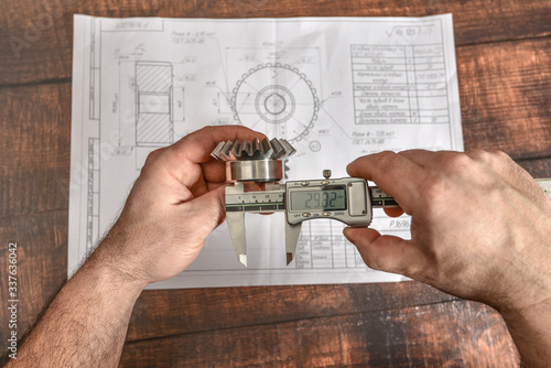 A working engineer checks the size of a bevel gear with an electronic vernier caliper against the background of a technical drawing.