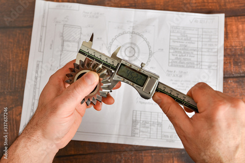 A working engineer checks the size of a bevel gear with an electronic vernier caliper against the background of a technical drawing.