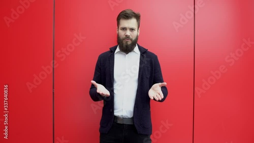 Young brunette bearded man is gesturing and talking something to the camera against red background. Guy in black jacket and white shirt is explaining or giving advice near red wall.