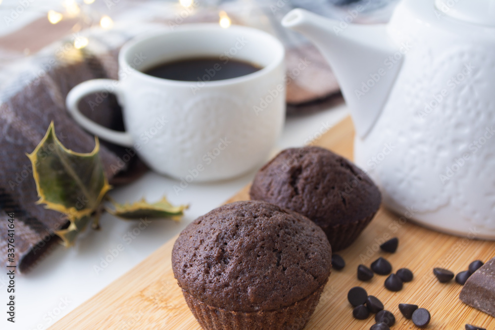 Chocolate muffins, chocolate chips, chocolate bar, a cup of coffee, teapot on a wooden surface with a warm scarf, Christmas lights bokeh and holly in the background Chocolate muffins, chocolate chips, chocolate bar, a cup of coffee, teapot on a wooden surface with a warm scarf, Christmas lights bokeh and holly in the background