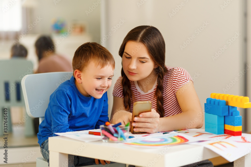 Motherhood concept. Brunette and son play with plastic building blocks. mom show somethimg funny in phone. Excited preschooler playing with his smiling mother or kindergarten teacher. Motherhood concept. Brunette and son play with plastic building blocks. mom show somethimg funny in phone. Excited preschooler playing with his smiling mother or kindergarten teacher.