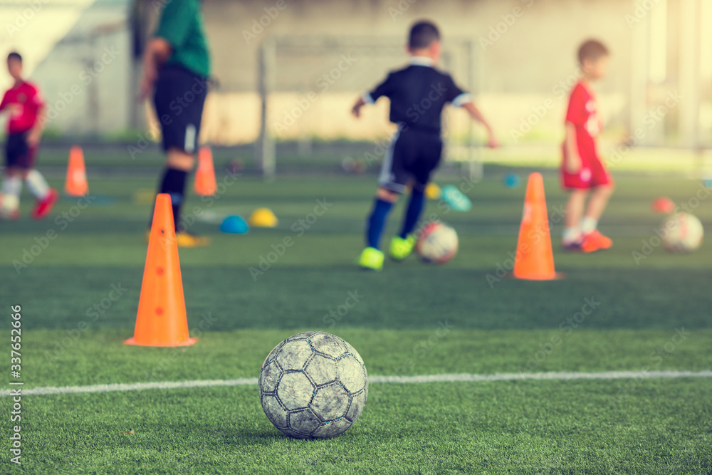 Old soccer ball on green artificial turf with blurry soccer team training. Blurry kid soccer player jogging between marker cones Old soccer ball on green artificial turf with blurry soccer team training. Blurry kid soccer player jogging between marker cones