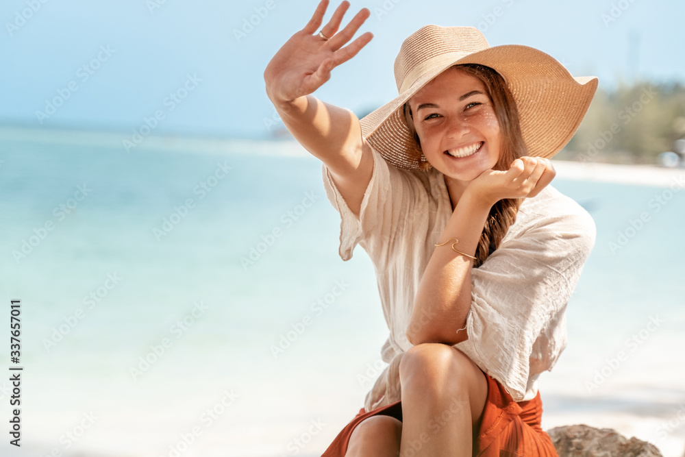 Beautiful young woman with long curly hair and a beautiful smile in a straw hat on a background of a turquoise tropical sea in a resort, travel and lifestyle concept