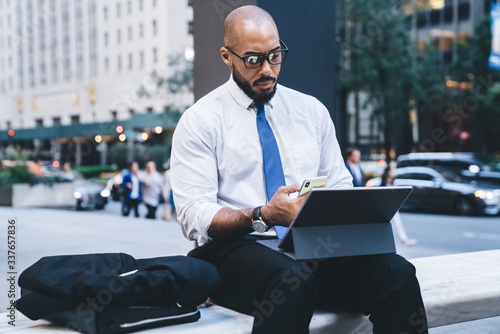 Black boss sitting with tablet and using smartphone