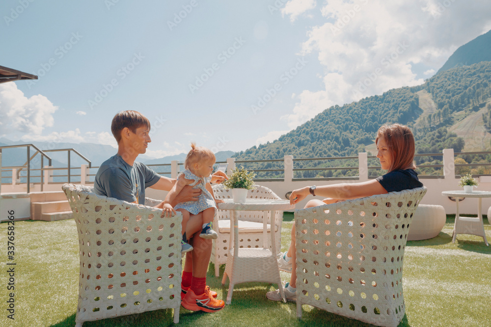 Beautiful young family, parents and little daughter, on the opened terrace in the white wicker furniture with great mountain view backwards, sunny day, blue sky - obrazy, fototapety, plakaty 